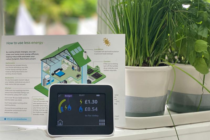 Smart meter dashboard showing electricity and gas energy consumption on kitchen window sill, next to herb plants. Laundry drying outside in background.