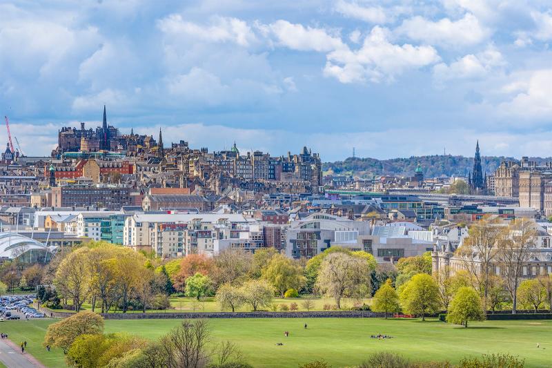 A cloudy blue sky over a sun-dappled Edinburgh Castle, with Holyrood Park, the Scottish Parliament, and Holyrood Palace visible in the foreground.