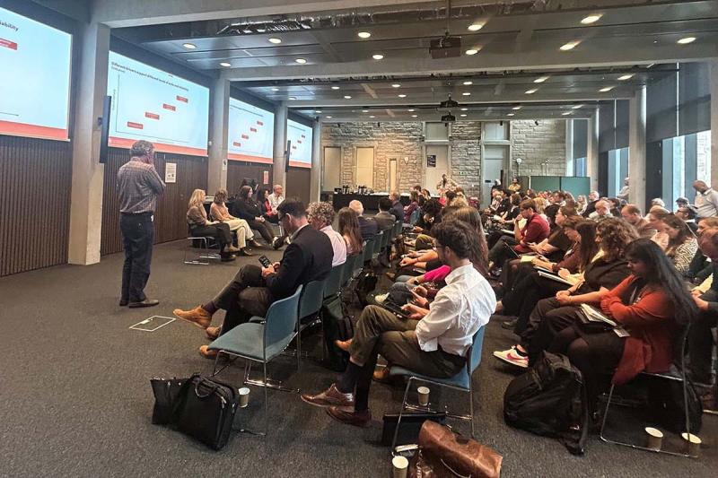 Side-on view of rows of attendees watching a presentation with speaker and panel