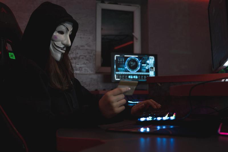Masked hacker with credit card at computer