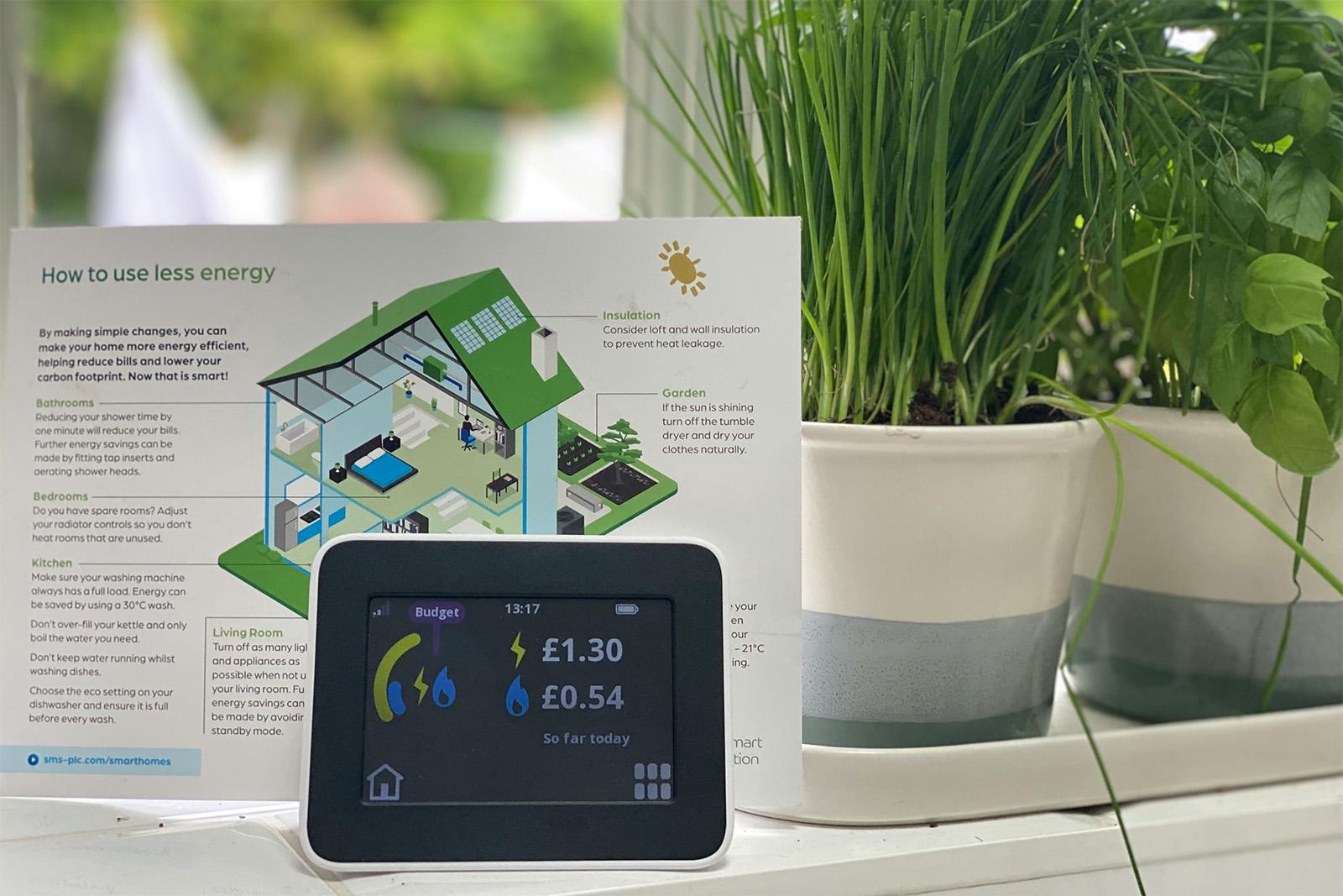 Smart meter dashboard showing electricity and gas energy consumption on kitchen window sill, next to herb plants. Laundry drying outside in background.