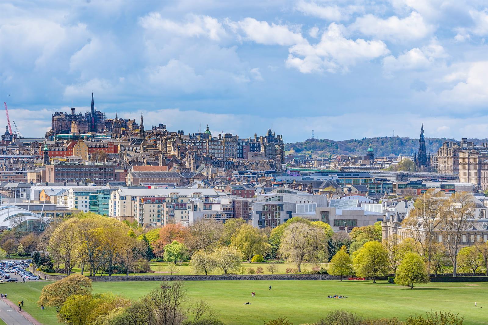 A cloudy blue sky over a sun-dappled Edinburgh Castle, with Holyrood Park, the Scottish Parliament, and Holyrood Palace visible in the foreground.